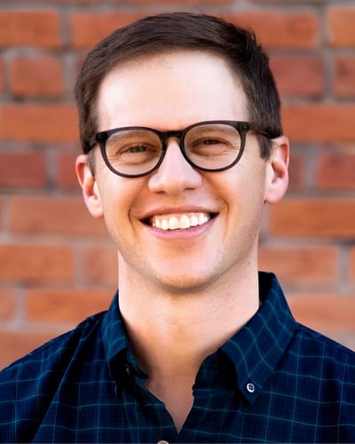 Adrian Chenault smiling, wearing round black-framed glasses and a dark teal plaid shirt, with a brick wall in the background